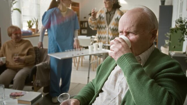 Senior man taking pill and grimacing in nursing home with people in background. Video for senior care and pharmaceutical marketing campaigns