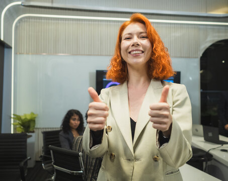 Confident Young Businesswoman . smile ginger-haired female professional blazer show thumbs-up in modern high-tech office representing corporate success positive results professional approval.