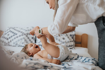 A mother plays with her laughing baby on a bed during a diaper change. The parent lifts the child's...