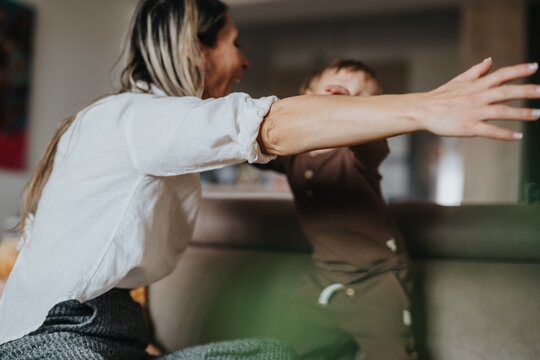 A mother reaches out to embrace her toddler on a living room couch. They share a playful, affectionate moment in a cozy home setting.