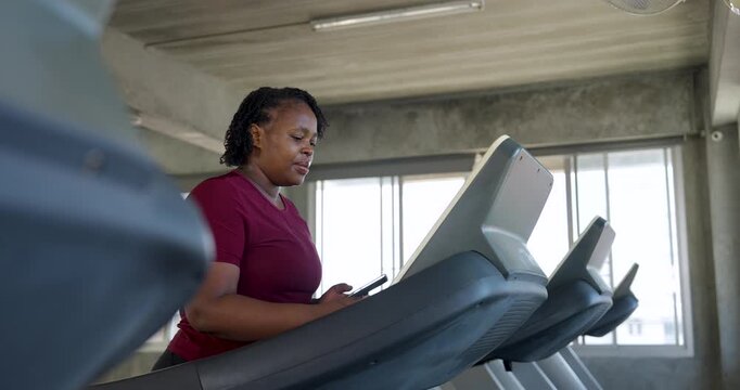 African American plus size woman jogging on treadmill, glancing at display while keeping steady pace. Curvy female athlete doing cardio workout for endurance, wellness, body confidence in gym.