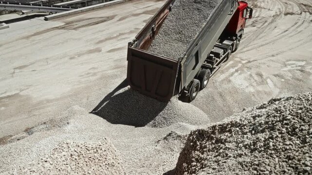 Industrial dump truck unloading crushed stone into aggregate yard under strong sunlight, elevated oblique view beside growing stone pile and dusty production area