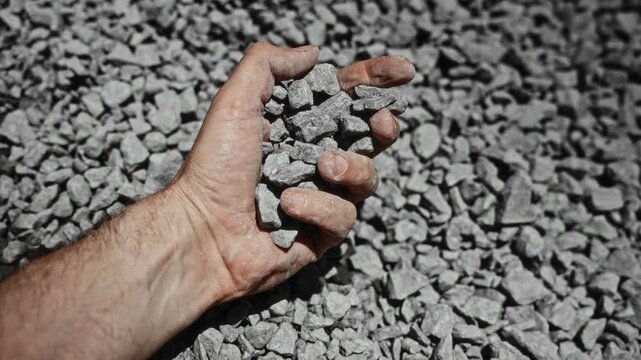 Adult male hand inspects crushed stone, squeezing and releasing handful over gravel bed in bright sunlight, showing rough texture, dust, angular fragments, and construction material detail