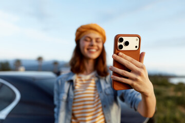 Fototapeta premium A cheerful woman takes a selfie outdoors, smiling at her phone in bright daylight. A casual denim jacket and striped shirt convey a relaxed, everyday lifestyle moment as the road and distant