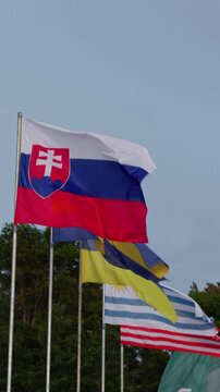 Slovak national flag waving on tall pole with other country flags behind it above treeline, open blue sky and soft daylight creating calm patriotic outdoor display