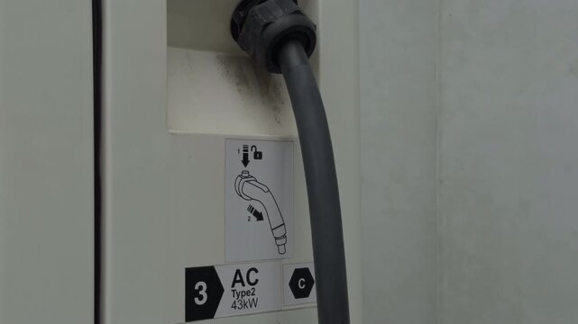 Close-up of electric vehicle charging connector plugged into public charging station with instruction label, dark cable, worn surface marks, and clean industrial detail for transport energy use