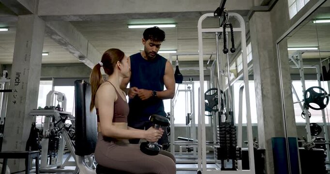 Young Asian couple during gym coaching session, woman seated with dumbbell while man demonstrates arm training cue, partner workout with support, motivation, healthy lifestyle, relationship teamwork.