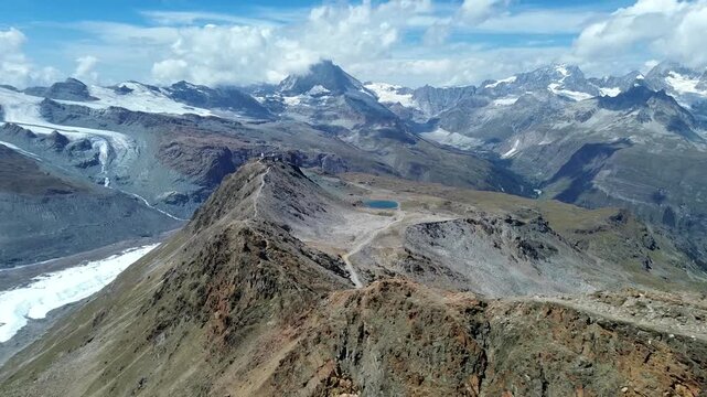 Aerial view of the Gornergrat ridge with rocky terrain, alpine glaciers, and distant snow-capped peaks under a blue sky in Zermatt, Valais, Switzerland.