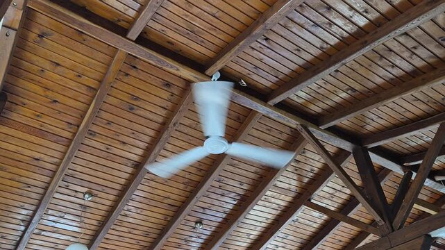 A spinning white ceiling fan is mounted on a rustic, vaulted wooden plank ceiling with heavy support beams