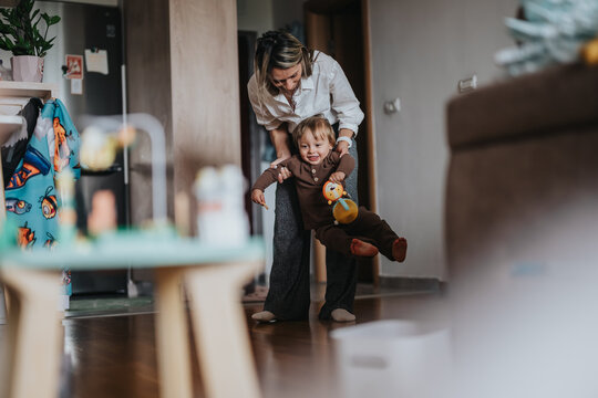 A mother plays with her toddler son in a cozy living room while lifting him and smiling. The joyful child clutches a small plush toy as the parent supports him during playful indoor time.