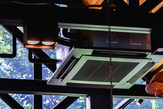 Modern cassette type air conditioner installed on loft ceiling with steel beams and wooden roof. Ceiling-mounted cassette air conditioning unit integrated into modern interior design. HVAC systems.