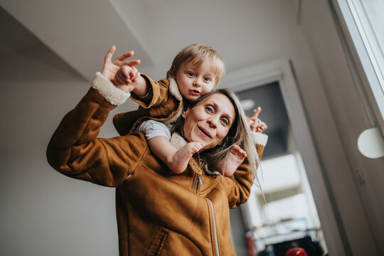 A mother carries her toddler on her shoulders indoors, both wearing warm jackets. The playful moment shows a close family bond and everyday life at home.