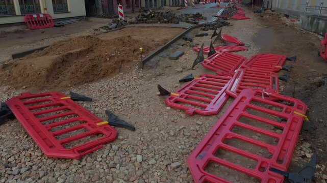 Road repair work with red safety barriers scattered beside open trench, broken cobblestone pavement, dirt piles, and temporary street closure setup in narrow urban construction zone