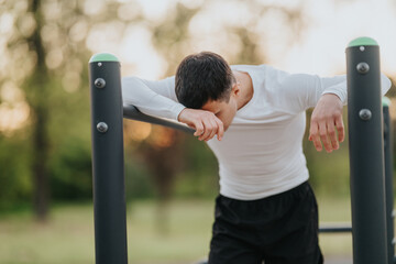 A young man rests his head and arms on an outdoor pull-up bar, exhausted from exercise. He leans on...