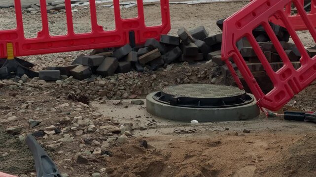 Construction barriers around utility access cover on rocky excavation site with scattered rubble, wet sand, and temporary roadwork safety setup during outdoor repair work