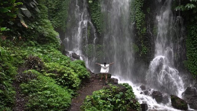 Young woman standing with arms outstretched enjoying the powerful flow of a tropical waterfall