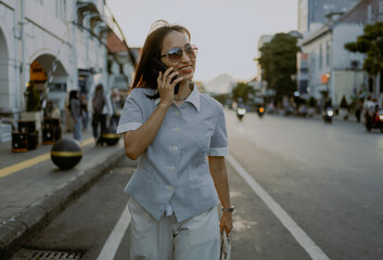 Fototapeta premium Fashionable style adult Asian Businesswoman commuting in sunglasses and casual button down shirt talking by phone call while walking in city at sunsets.