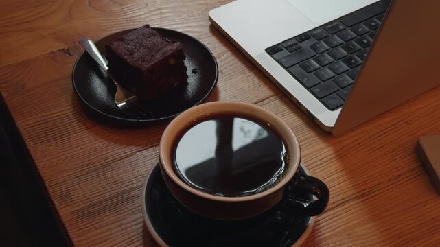 Coffee cup beside chocolate brownie and laptop on wooden desk, creating cozy remote work break scene with warm window light, overhead composition and relaxed home office mood