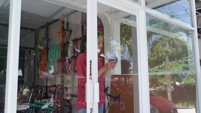 Woman near left side opens and adjusts glass sliding door at home entrance, with indoor bicycle, red curtain, and bright tropical reflections shifting across