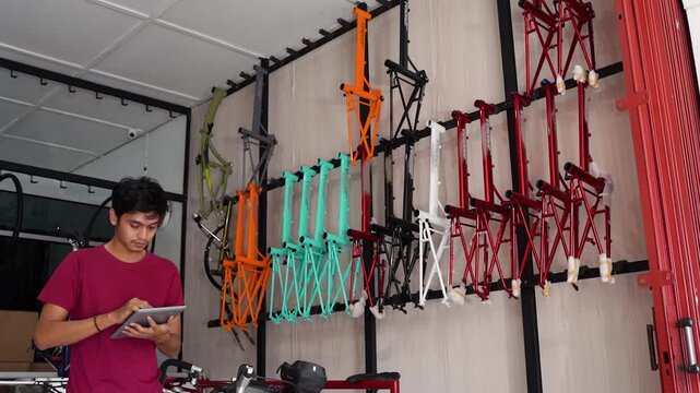 Man stands at left using tablet inside bicycle workshop, while colorful frames hang on wall behind him and repair tools mark organized industrial workspace