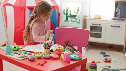 Naklejka premium Little girl drawing with crayon at red table surrounded by toys in a playroom building; creative playfulness.
