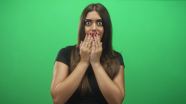 Woman covers mouth with hands, wide eyes and burgundy painted nails facing camera against green studio backdrop; surprise curiosity.