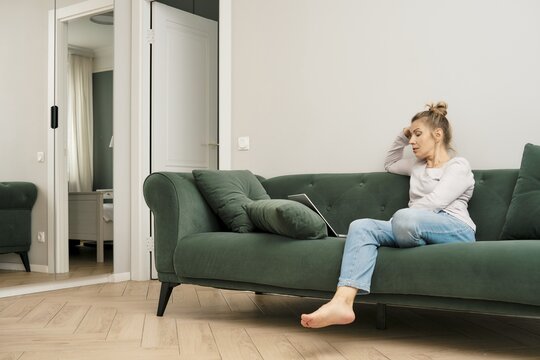 Middle-aged woman sits cross-legged on a green couch, focused on her laptop. The modern living room features minimalistic decor, with a mirror reflecting natural light and a doorway in the background