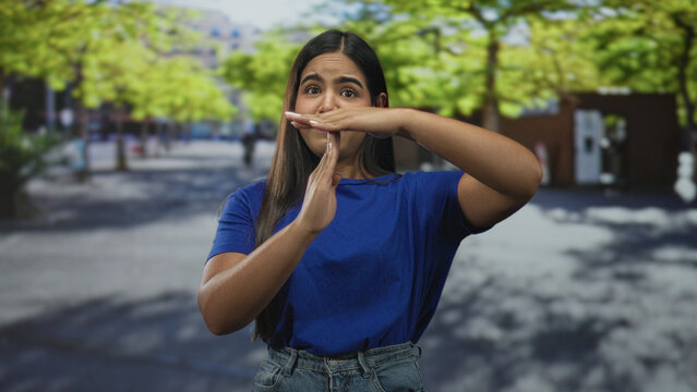Young hispanic woman forms timeout gesture with hands and forearms visible while standing in a tree lined urban street plaza wearing blue shirt and jeans; assertive pause.