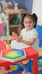 Obraz premium Girl drawing with yellow pencil at a red children's table, colored pencils cup and rainbow wooden stacking toy visible in preschool building; concentration creativity.