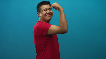 Fototapeta premium Hispanic man flexes bicep and smiles wearing red t shirt, side pose in studio with teal backdrop; confidence strength.