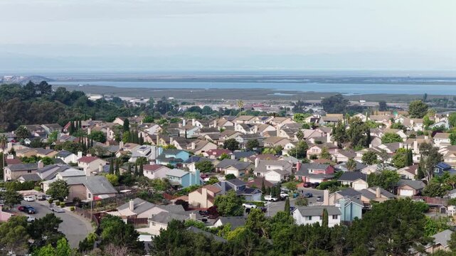 Single-family homes with mixed rooftops and colorful facades spreading across hillside toward San Pablo Bay and tidal wetlands under hazy sky in Vallejo, California, USA. Aerial drone panorama shot