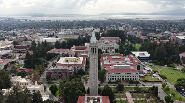 UC Berkeley campus with Sather Tower rising above red-roofed academic buildings and lush grounds with San Francisco Bay stretching to horizon in Berkeley, California, USA. Aerial drone approaching