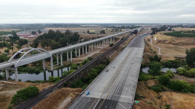 High-speed rail viaduct with arched river bridge runs parallel to a freeway carrying moving traffic across flat Central Valley farmland in Fresno County, California, USA. Aerial drone wide shot