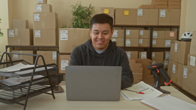 Man at laptop smiling and typing while glancing at phone amid stacked parcels in a building; small business contentment.