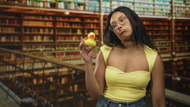 Woman holding rubber duck, pouting lips and lifting it up for inspection while wearing yellow top and glasses in a building; boredom reflection.