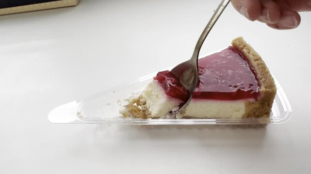 Slice of cheesecake with red jelly topping rests on a clear plastic plate, as a fork prepares to cut into the dessert, revealing its creamy texture and crumbly crust in a well-lit indoor space