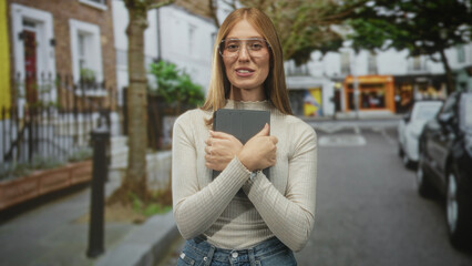 Fototapeta premium Young woman hugs tablet to chest with crossed arms on a street in front of shops; comfort confidence.