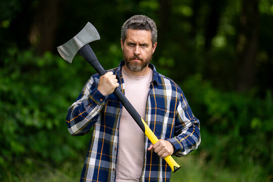 Bearded lumberjack. Lumberjack brutal bearded man in checkered shirt and ax in forest. Serious lumberjack holding axe and looking away outdoors. Lumberjack worker.