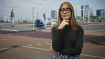 Fototapeta premium Woman with hand on chin and palm slightly raised at street intersection wearing glasses and black top; subtle contemplation.