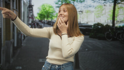 Fototapeta premium Woman points finger outward on street, hand to chin and visible pointing gesture, redhead in sweater and jeans; curiosity discovery.