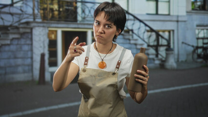 Fototapeta premium Woman in apron holding pottery bottle with finger pointing gesture toward camera in street; warning annoyed craft.