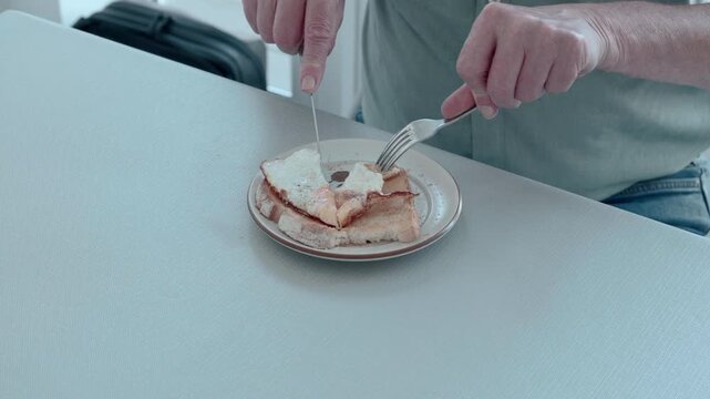 Man cuts toast with knife and fork at table. Hands steady, focused on simple meal. Quiet breakfast moment, calm, unhurried. Ideal for health, comfort, or morning routine scenes