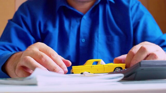 Optical zoom on car insurance agent  playing with yellow toy car on office desk. Close up, shallow depth of field, natural light. Businessman and miniature auto.