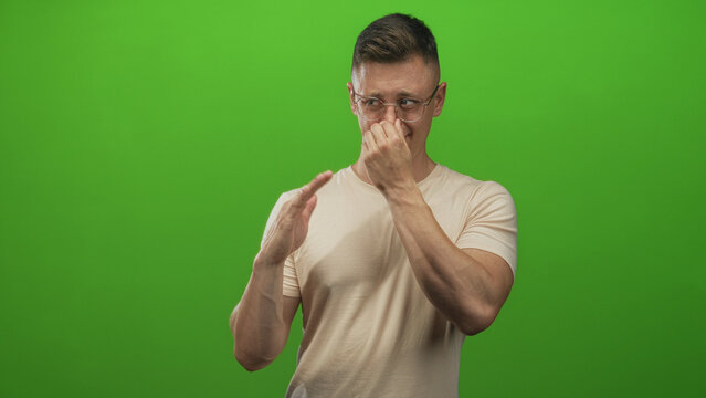 Man pinching nose with right hand and gesturing with left hand while grimacing in a green studio wearing glasses and beige shirt; disgust.