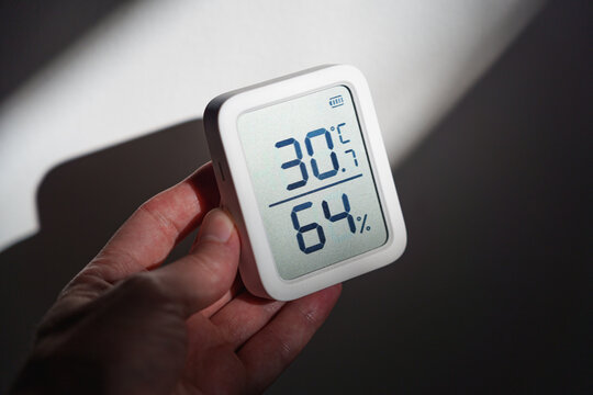 Hand holding digital thermometer showing 30 degrees Celsius indoors during a summer heat wave