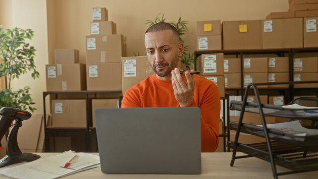 Man holds smartphone near mouth while seated at laptop with stacked cardboard boxes and packing supplies in building; small business focus.