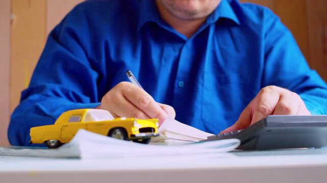 Close-up of an insurance agent filling out paperwork at his desk. Focus on his hands and pen, with a yellow toy car in the foreground. Shallow depth of field, natural lighting. 