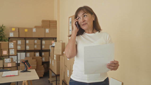 Woman holds phone to ear and reads invoice with hand while surrounded by cardboard boxes in a building; focused small business.