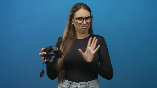 Woman holding dslr camera with raised hand and grimacing face showing disgust while checking lens in studio; disgust rejection.