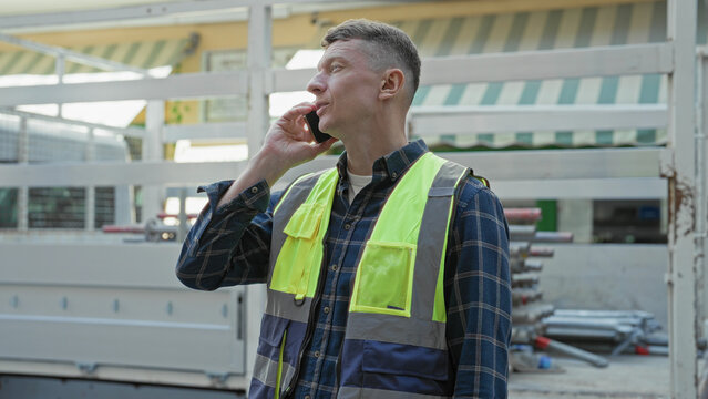 Man in yellow hi vis vest and plaid shirt holding smartphone to ear, smiling and looking up beside scaffolding and stacked metal pipes at building site; confidence.